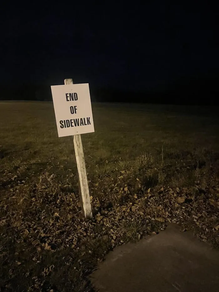 An unsettling liminal space: a white 'END OF SIDEWALK' sign in a dark, grassy field at night.