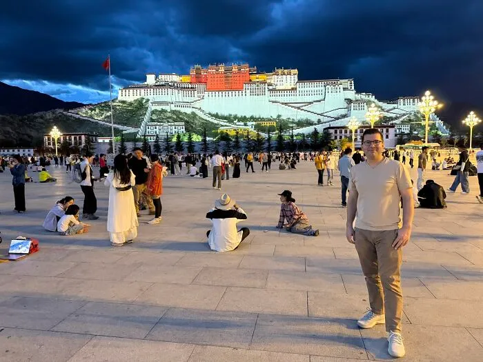 A traveler in China standing in a bustling plaza at dusk, with the illuminated Potala Palace in the background.