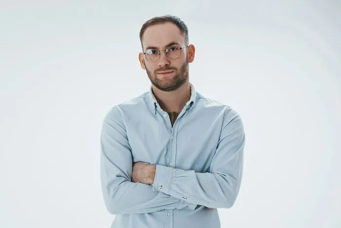 A secretly smart man in glasses with arms crossed, looking intently, showing hints of intelligence.