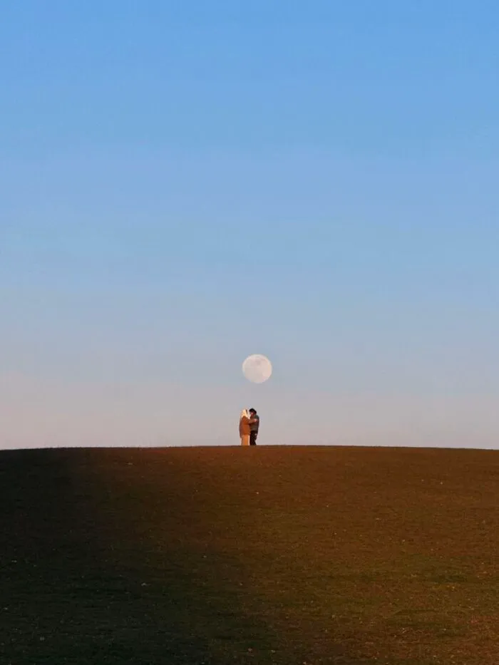 A couple stands on a hill, silhouetted against a large full moon, representing a wholesome photo trend.