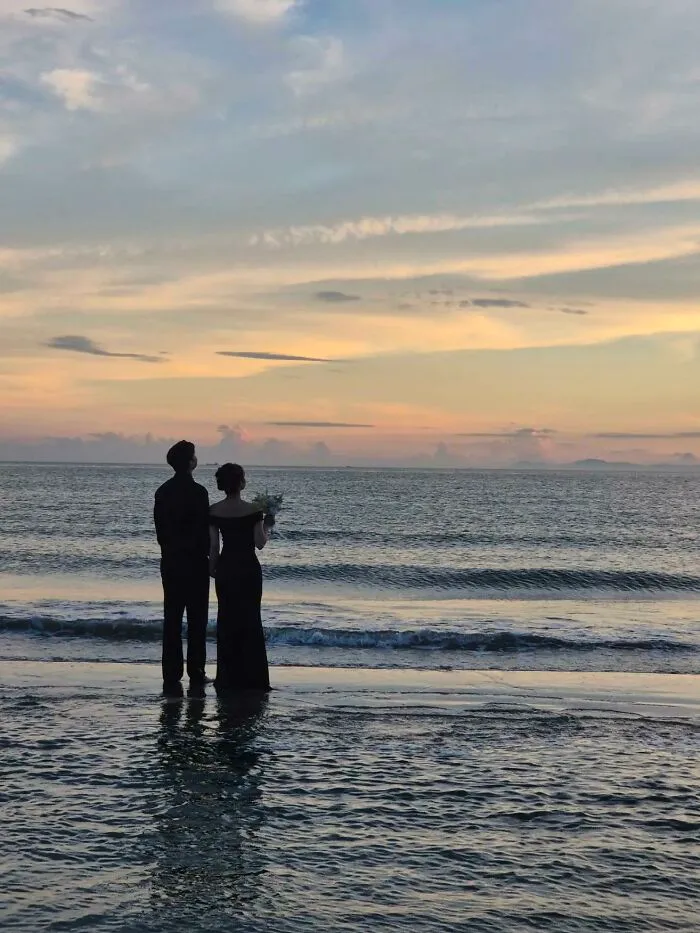 A couple in formal attire stands at the edge of the ocean, looking out at the sunset, embodying a wholesome photo trend.