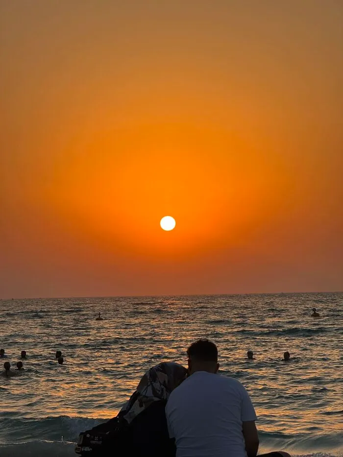 A couple watching the sunset over the ocean, like a painting, embodying the wholesome photo trend between strangers.