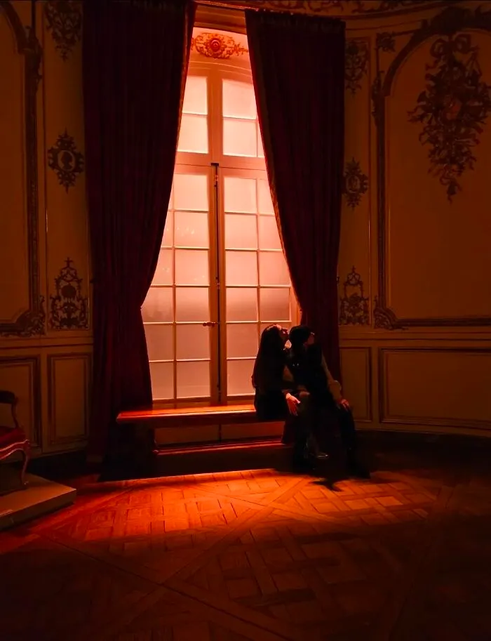 Two strangers sit by a window with red curtains, bathed in warm light, exemplifying the wholesome photo trend.
