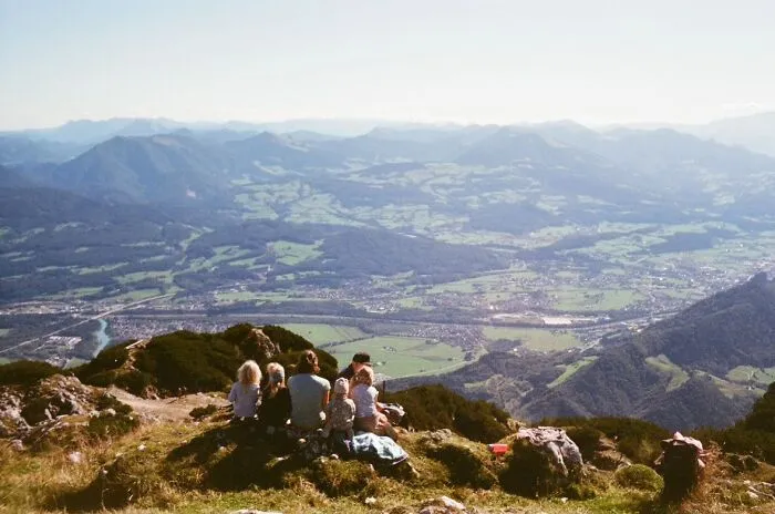 A group of people sit on a mountain peak, gazing at a vast landscape below, embodying a wholesome photo trend.