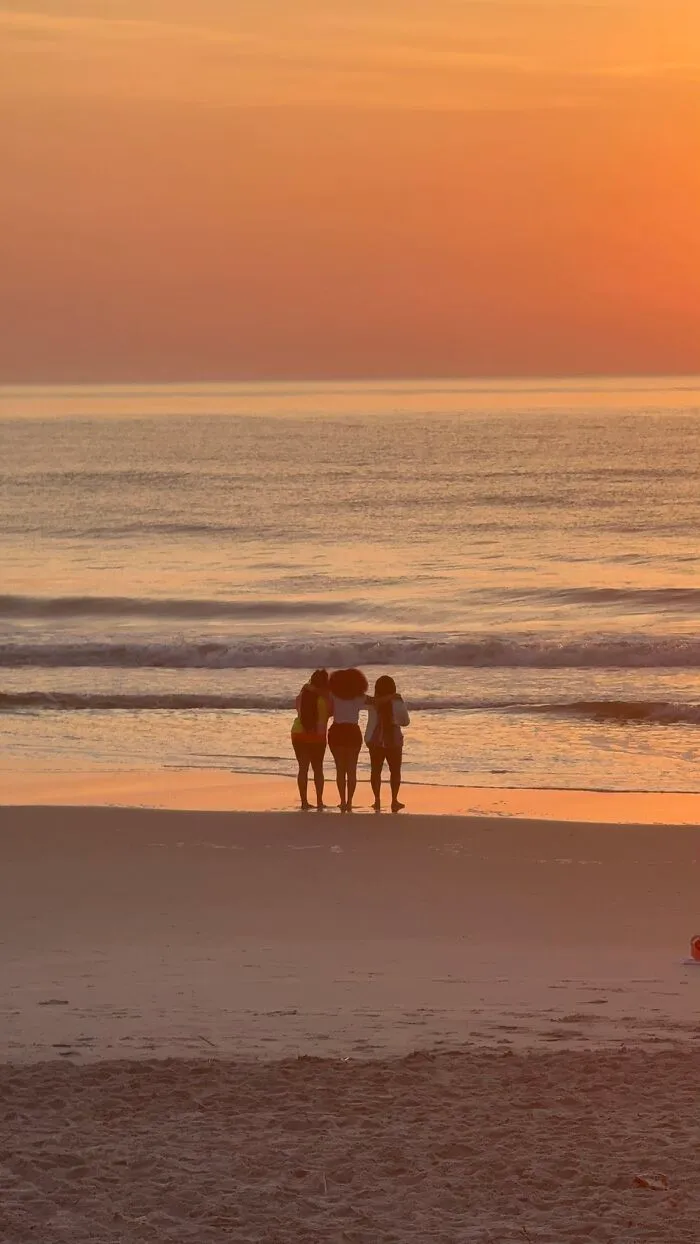 A wholesome photo trend featuring three friends standing together on a beach, silhouetted against an orange sunset.