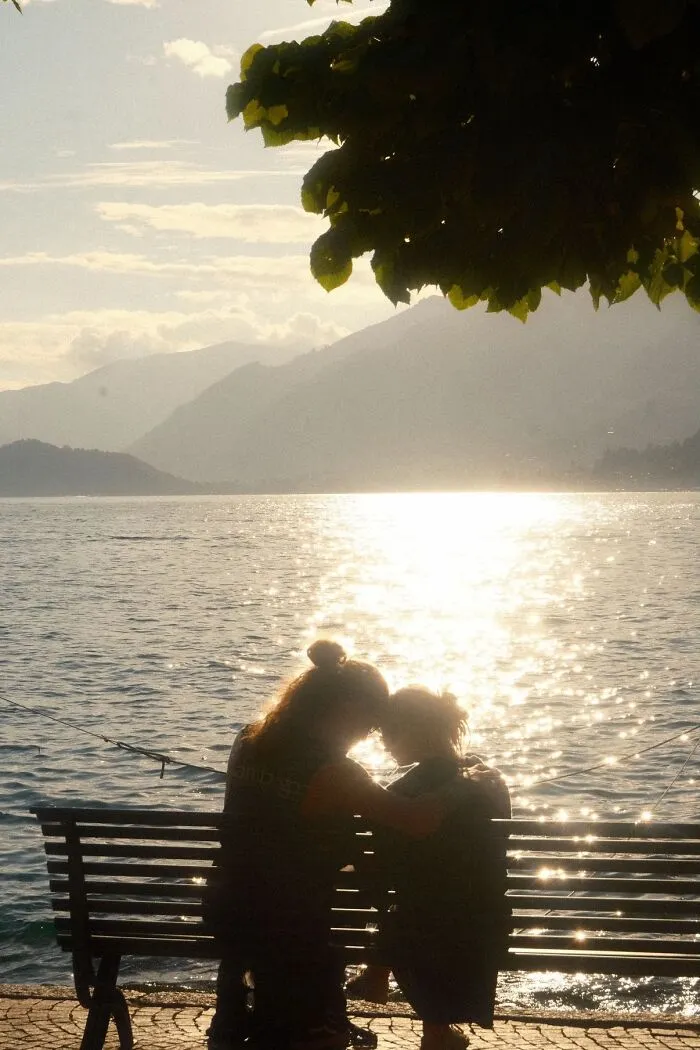 A wholesome photo trend of two people embracing on a bench by a shimmering lake with mountains in the background.