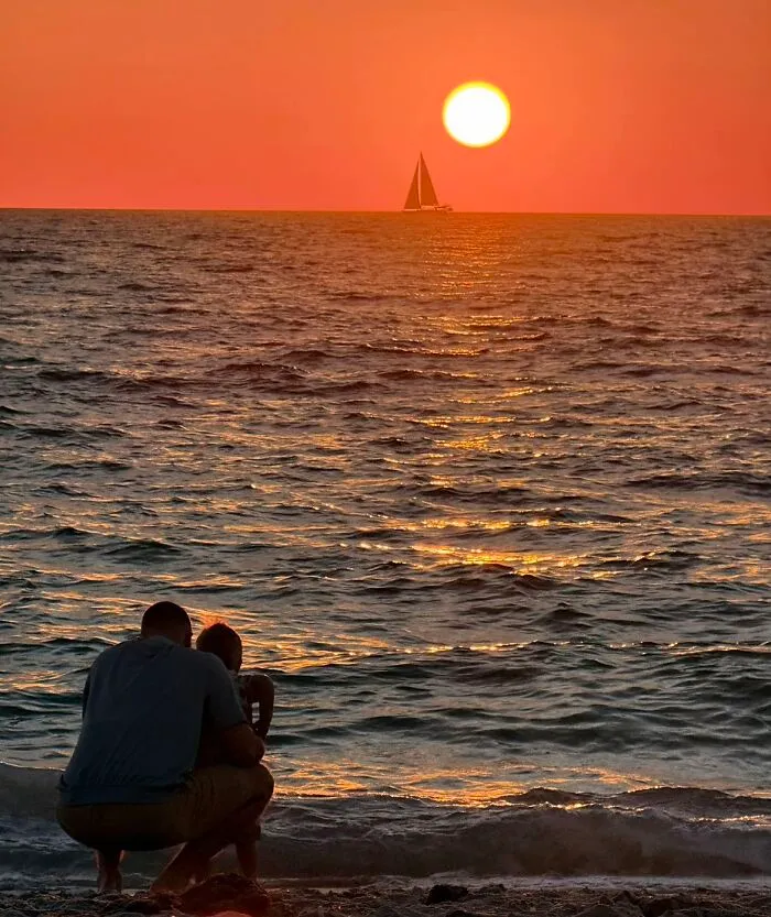 A wholesome photo trend showing a father and child watching a beautiful ocean sunset with a sailboat.