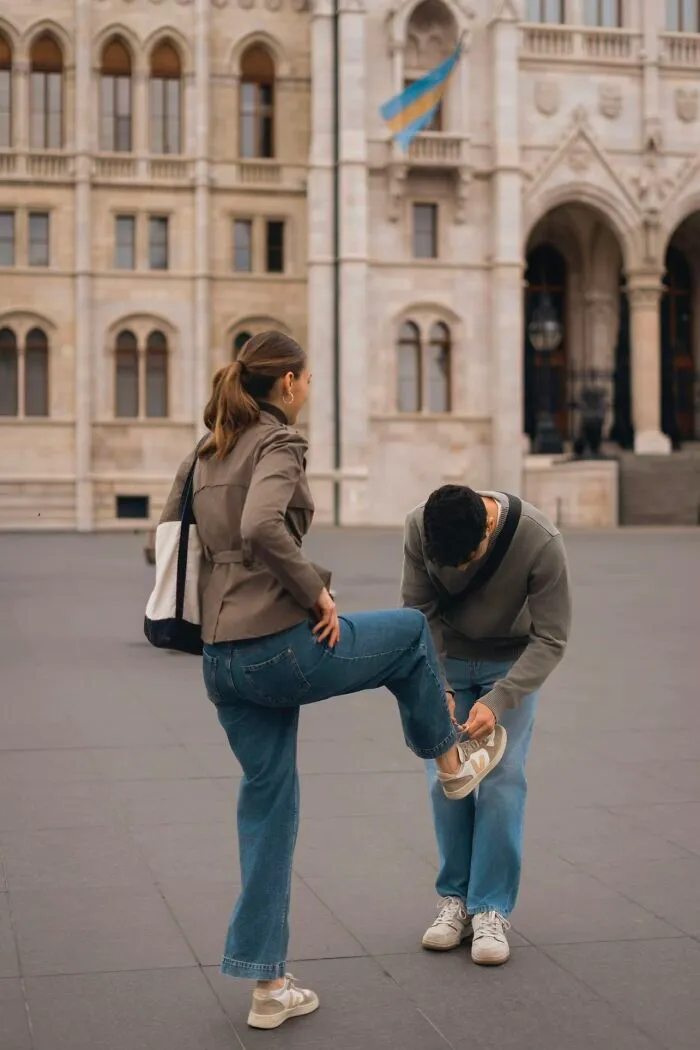 A man tying a woman's shoe in front of a grand building, a wholesome photo trend in a public space.