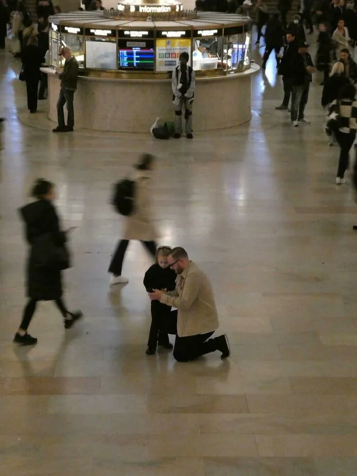 A father kneels to interact with his child in a busy station, a painting-like wholesome photo moment.