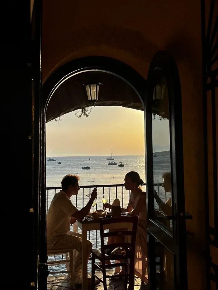 A couple dining on a balcony overlooking the sea at sunset, a serene scene from the wholesome photo trend.