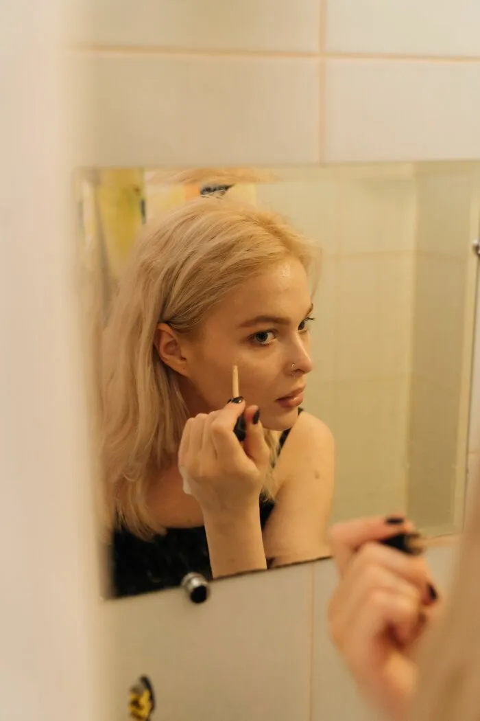A blonde woman in a mirror applies makeup, highlighting a beauty routine that might be a turn off for some.