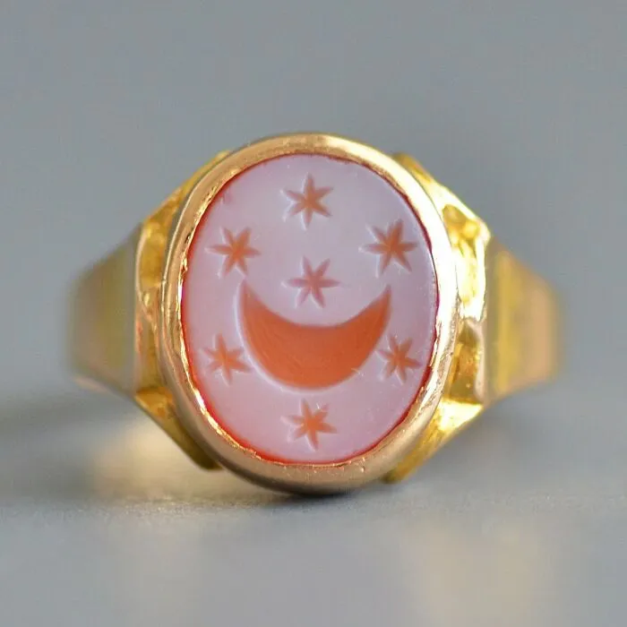 Intact after years, a gold ring featuring a pinkish-white oval stone engraved with a crescent moon and stars.