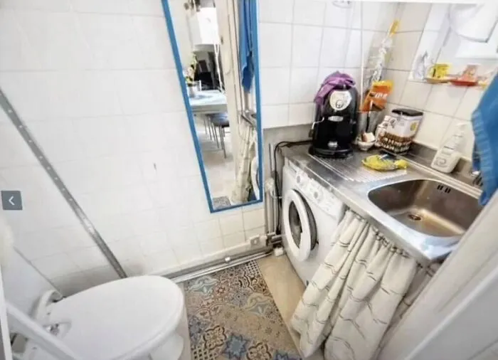 A cramped bathroom featuring a toilet, a washing machine integrated with a kitchen sink, reflecting unhinged real estate listings.