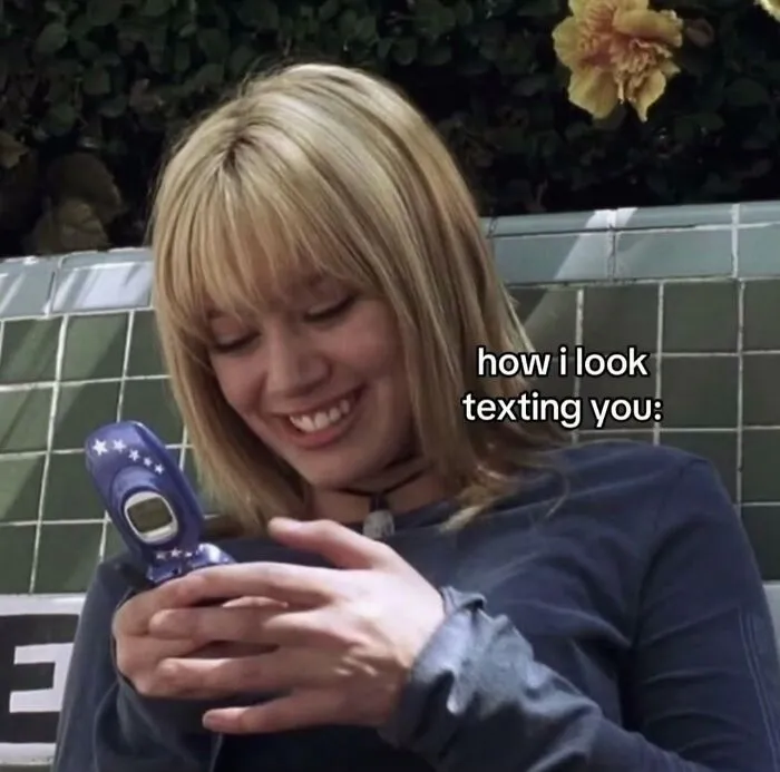 A relatable meme of a smiling woman texting on a flip phone captioned: how i look texting you.