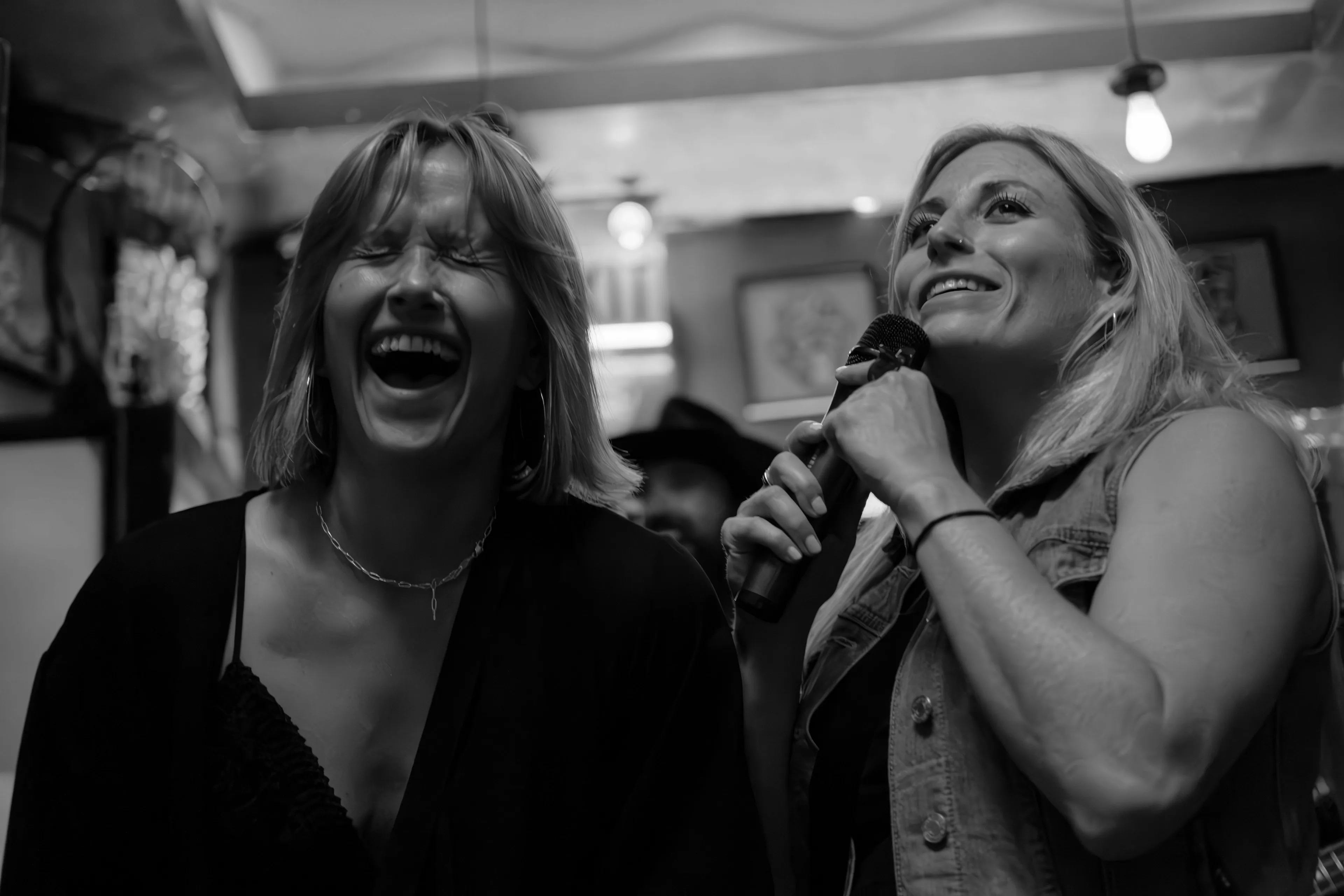 Two women enjoying karaoke night, one laughing heartily and the other singing into a microphone.