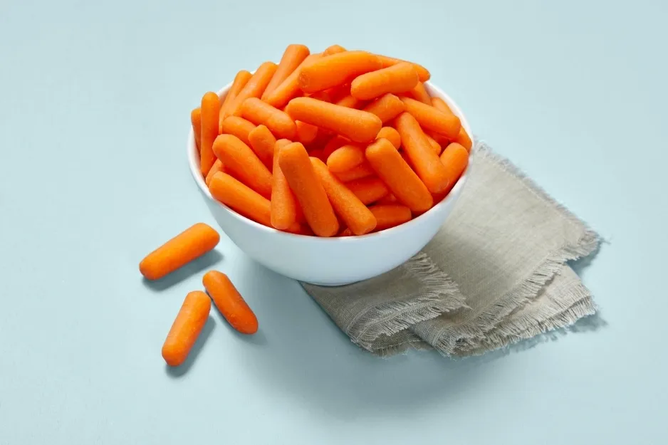 A white bowl filled with baby carrots, a healthy snack for a road trip, with some carrots scattered on a blue surface.