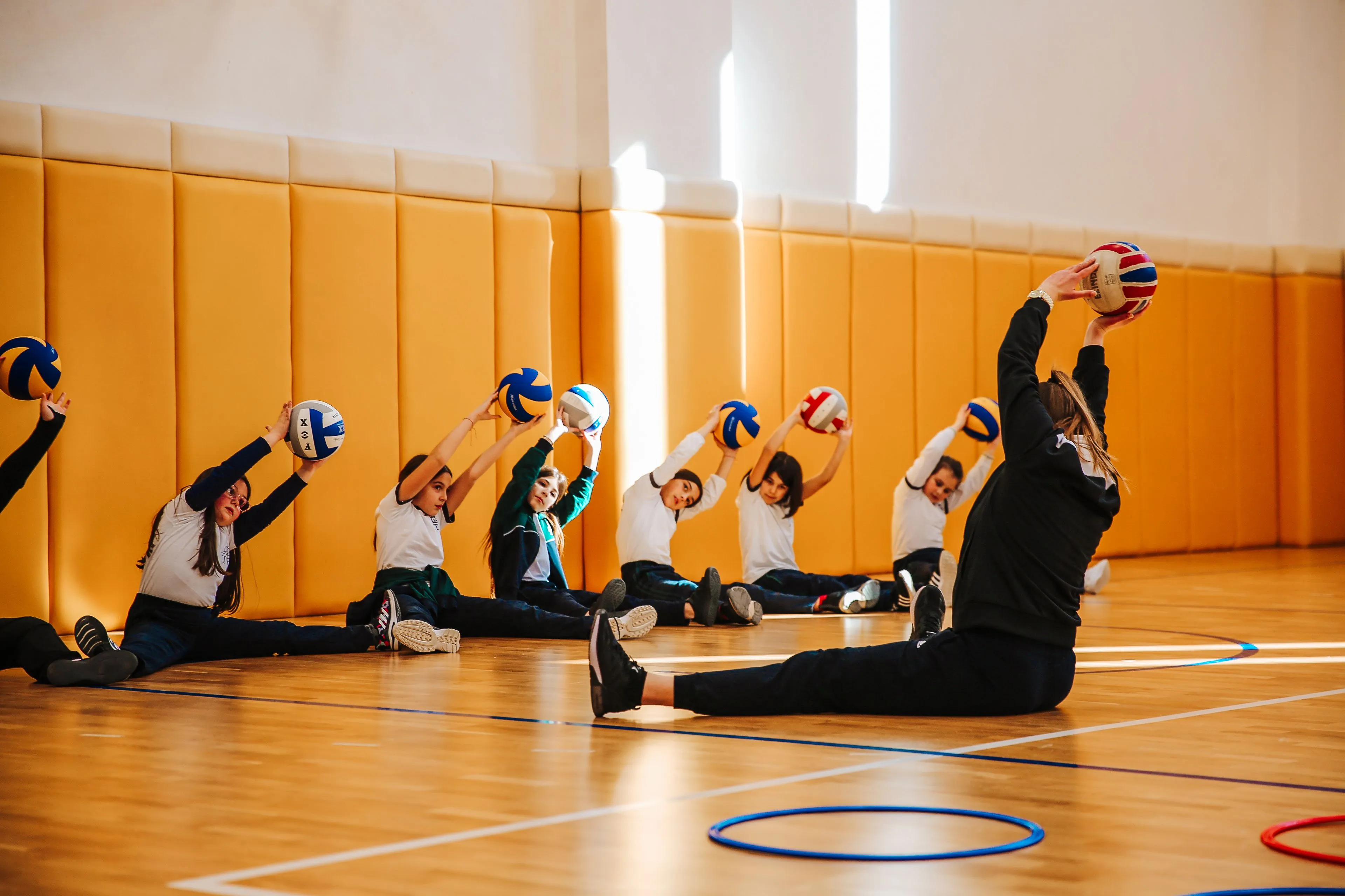 Children in a school gym doing stretches with balls, enjoying a PE class.
