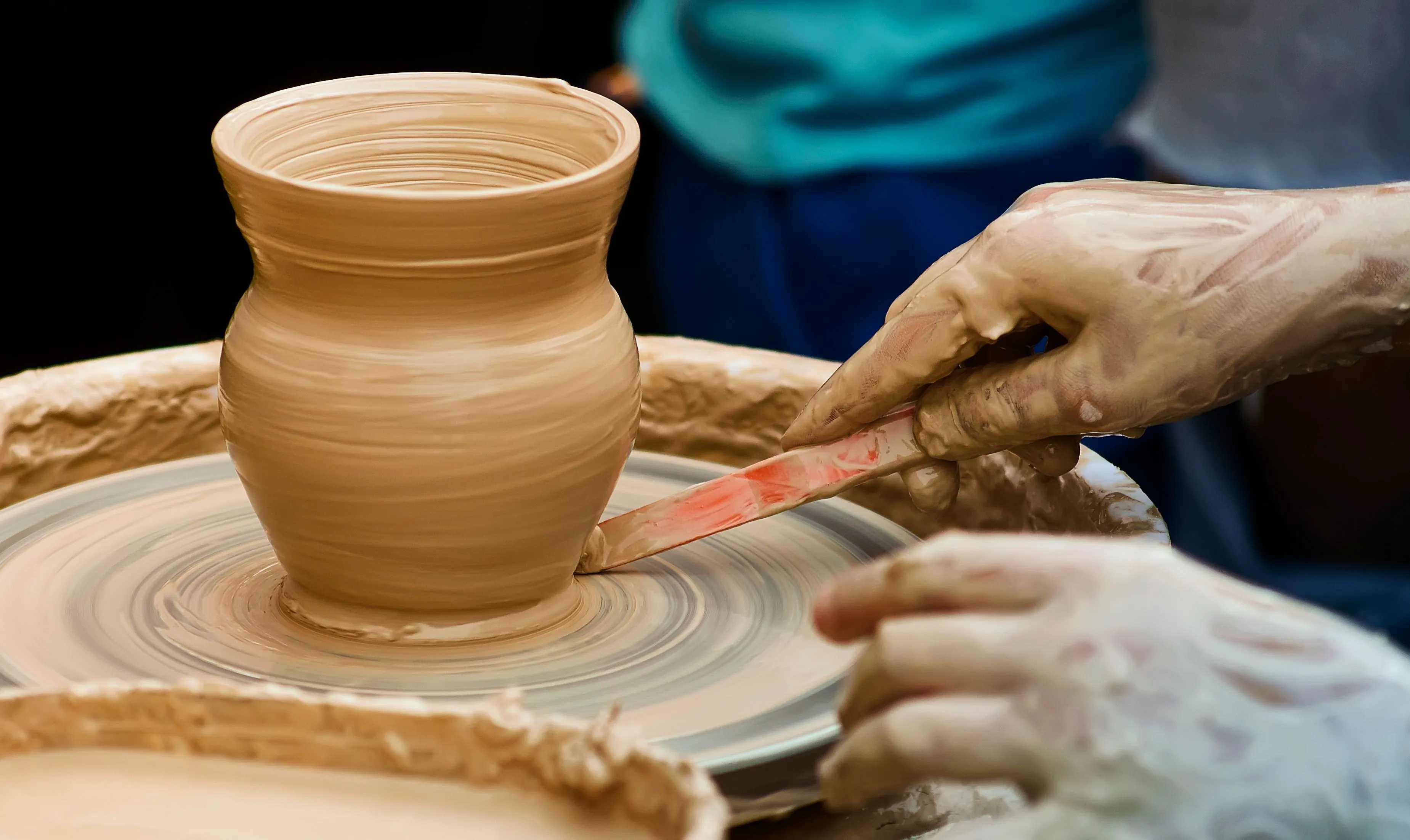 Hands covered in clay shaping a pot on a pottery wheel, representing the hobby of pottery.