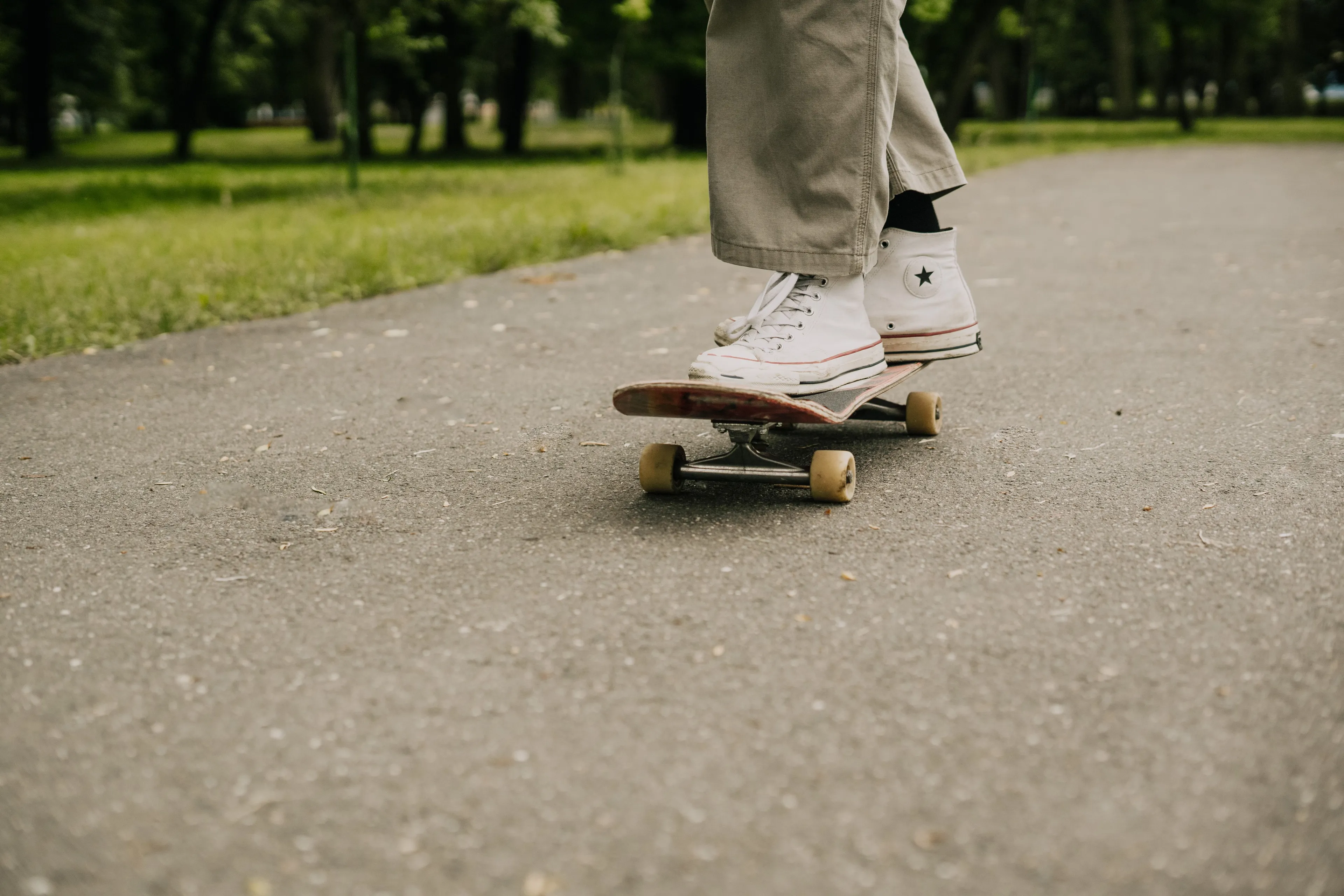A person's feet in white high-top sneakers stand on a skateboard on an asphalt path in a park, showing skateboarding.