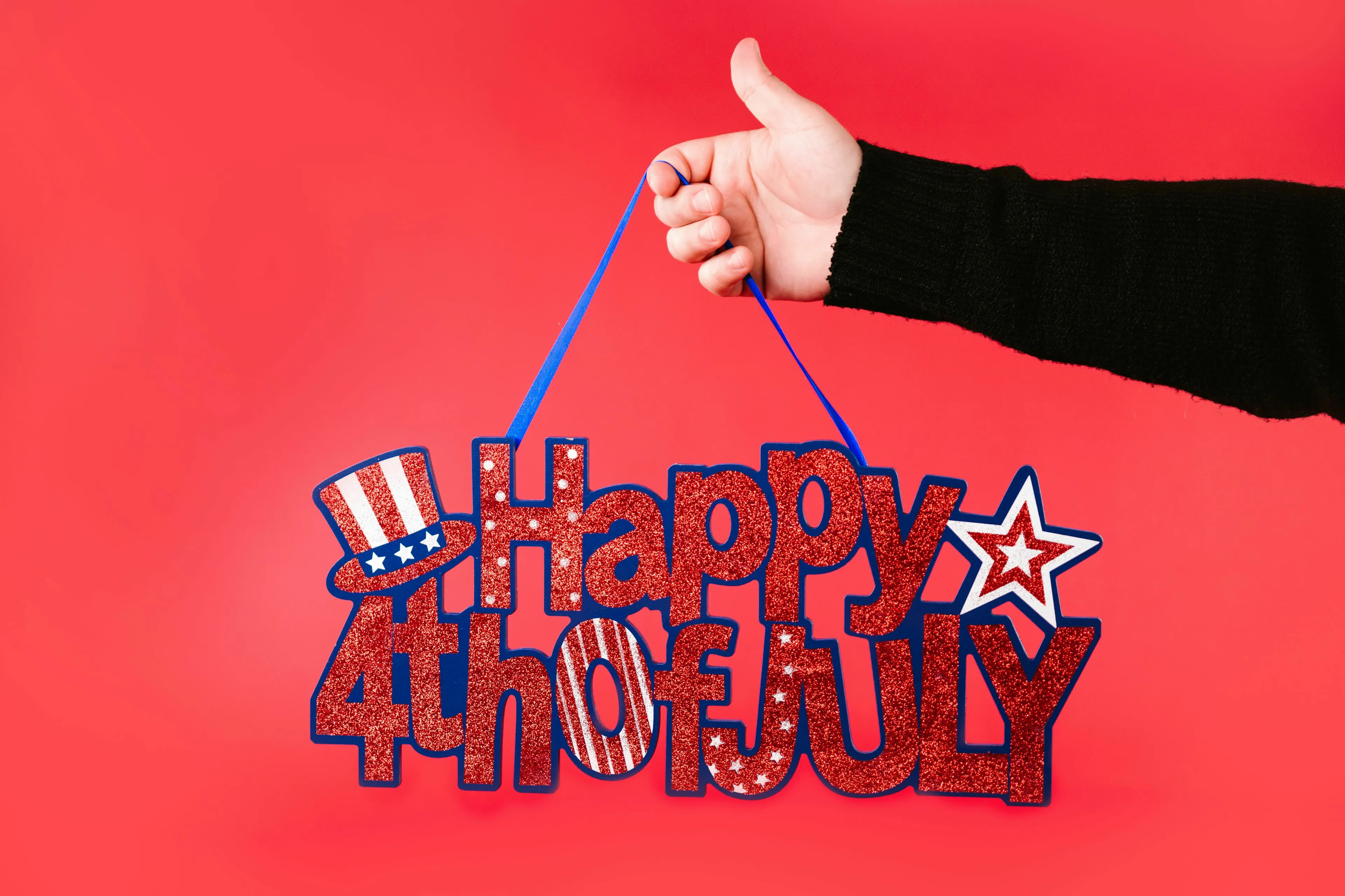 A hand holds a sparkly Happy 4th of July sign against a red background, celebrating the U.S. holiday.