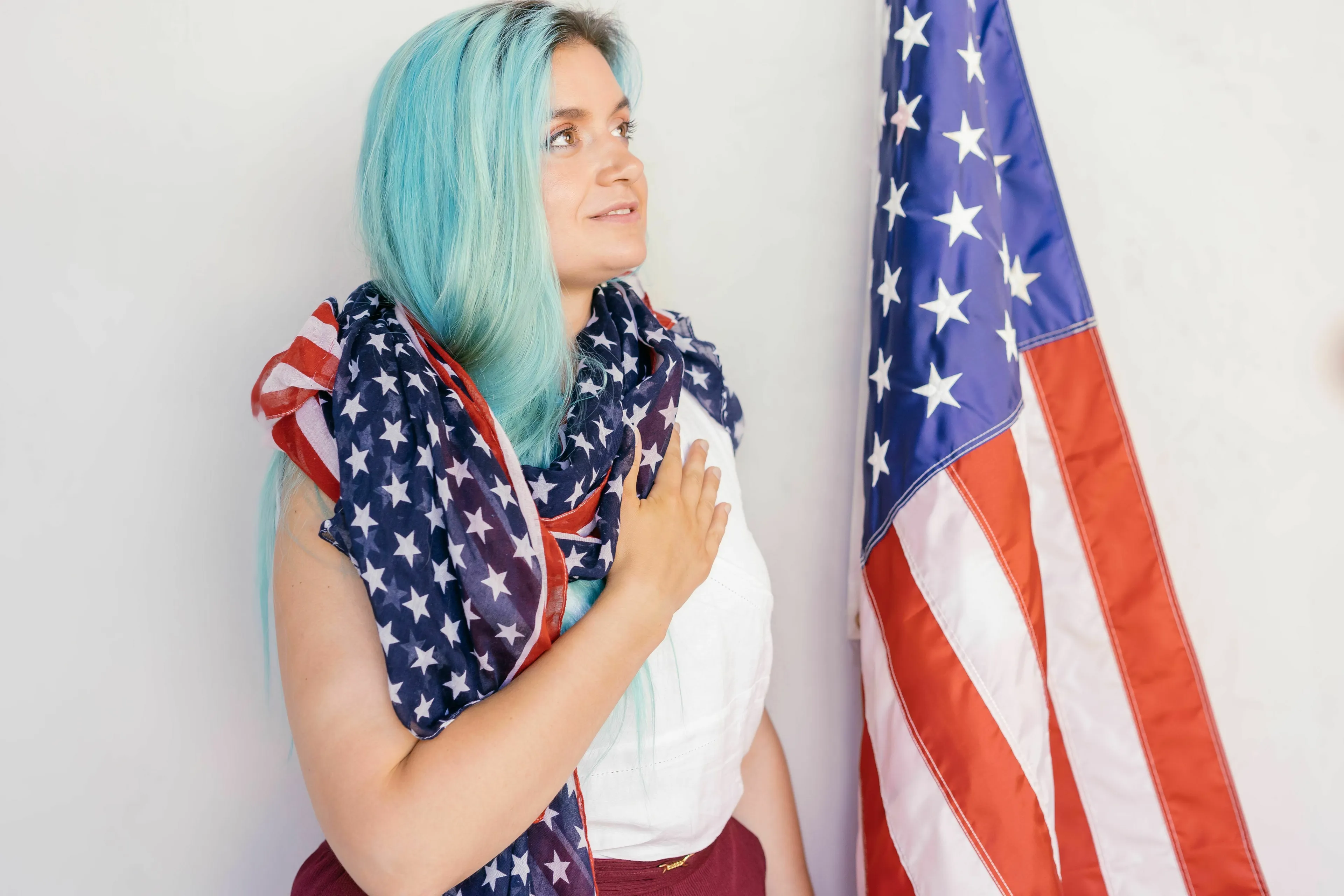A woman with blue hair, draped in an American flag, standing next to a flagpole, honoring the patriotic song and national anthem.