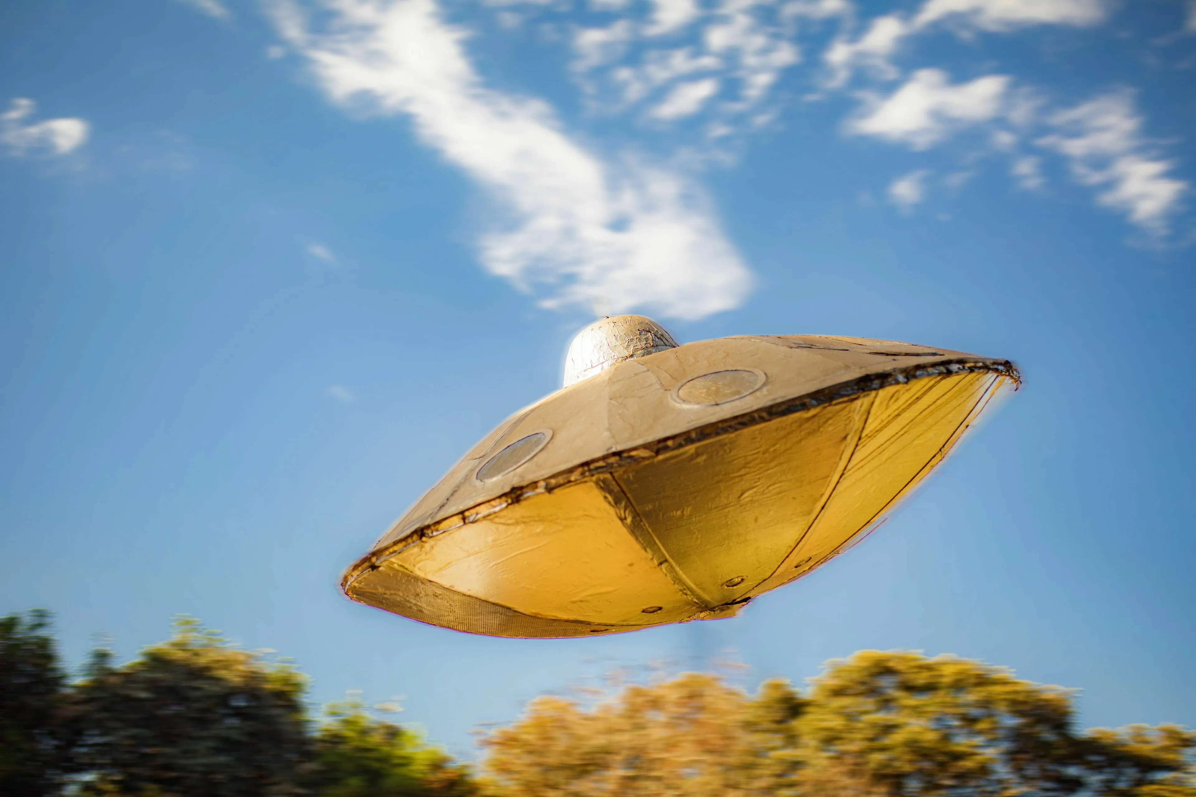 A gold-colored unidentified flying object (UFO) hovering against a blue sky with clouds.