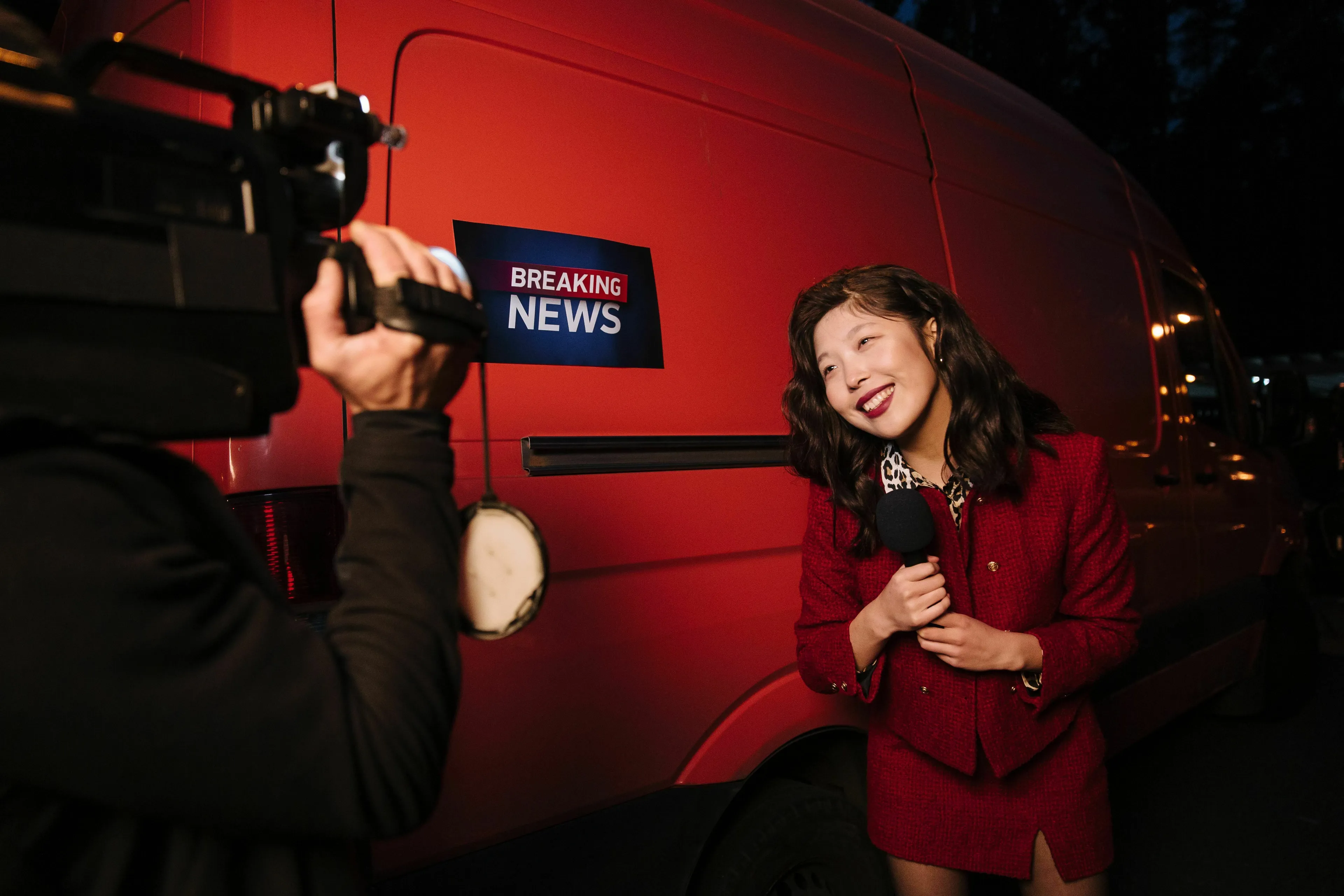 A smiling reporter in a red suit holding a microphone, being filmed in front of a breaking news van.