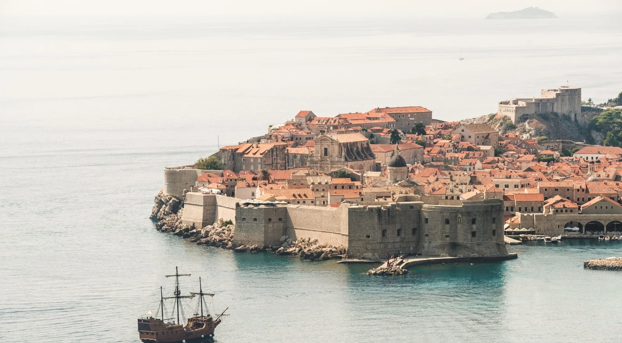 A historical European city with fortified walls along the coast, red-tiled roofs, and a tall ship on the water. General knowledge on Europe.