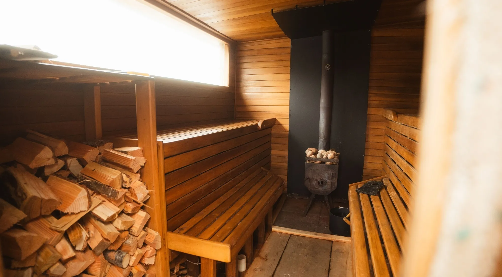 Interior of a traditional sauna with wooden benches, a stove with rocks, and a pile of firewood. European country.