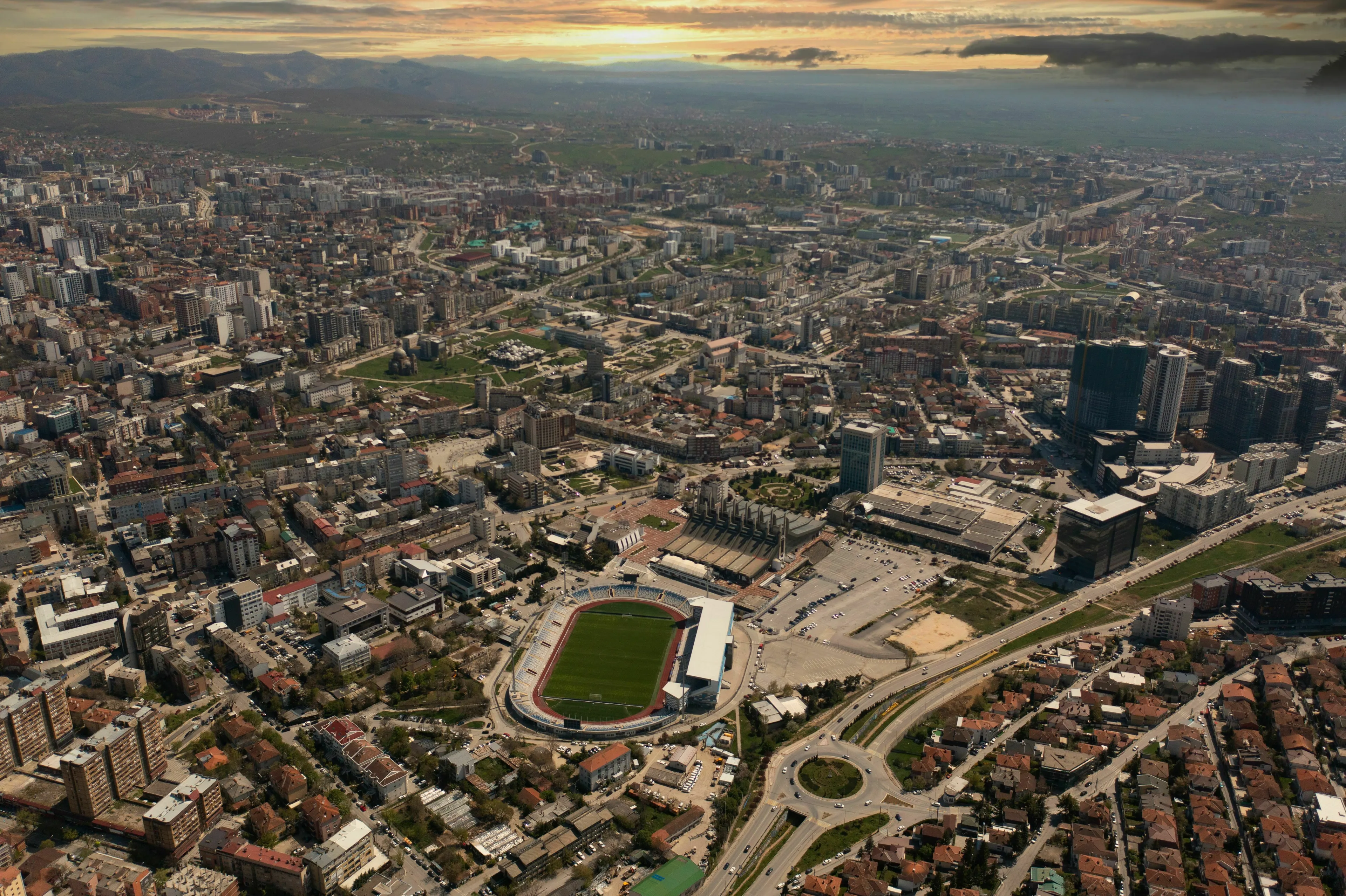 Aerial view of Pristina cityscape, featuring a large stadium and numerous buildings under a dramatic sky.