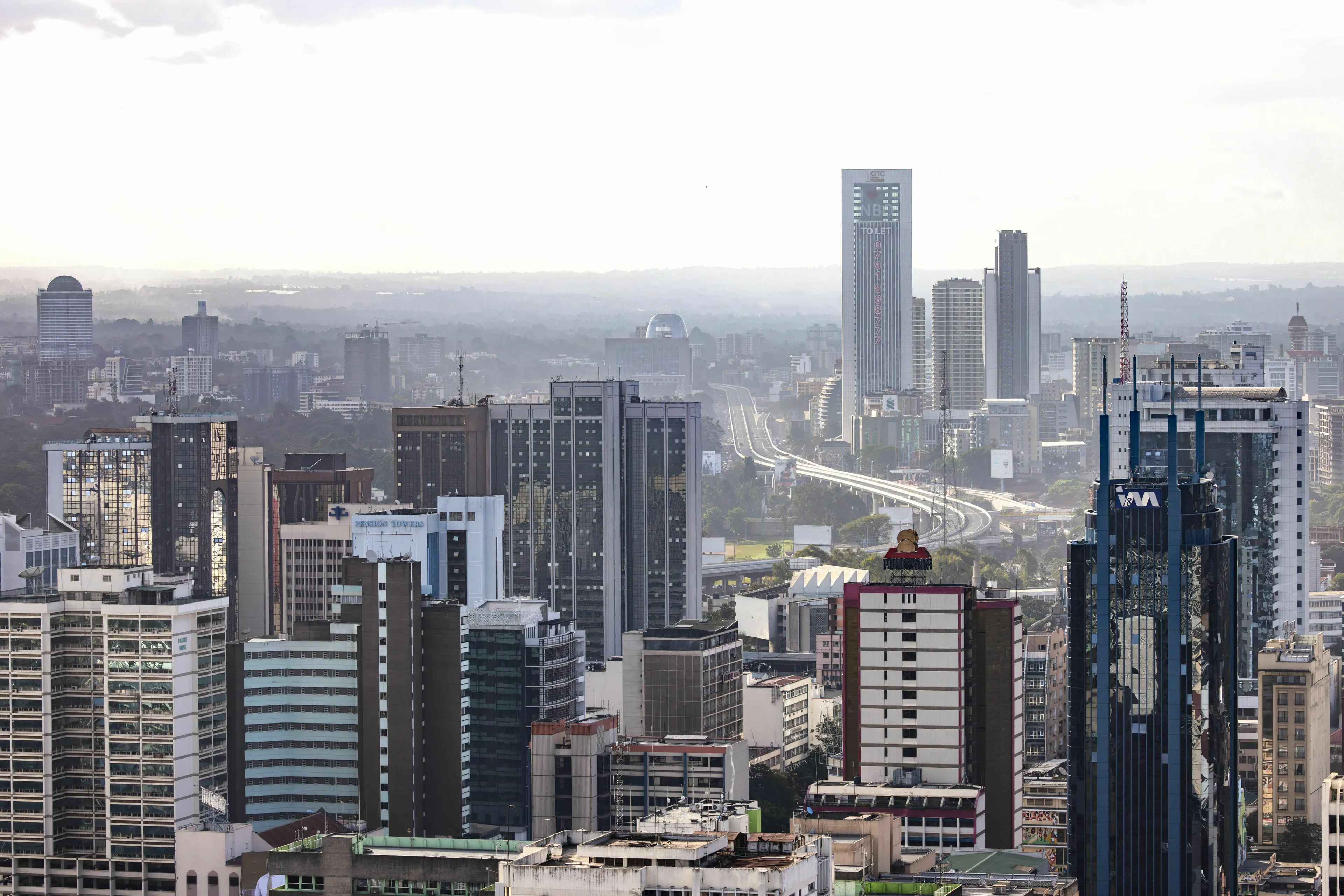 A sprawling cityscape of Nairobi with modern skyscrapers and a highway system visible against a hazy sky.