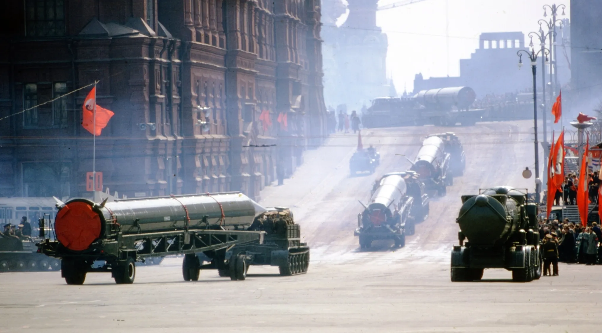A military parade showcasing missiles, central to the Cuban Missile Crisis, along a wide street with red flags.