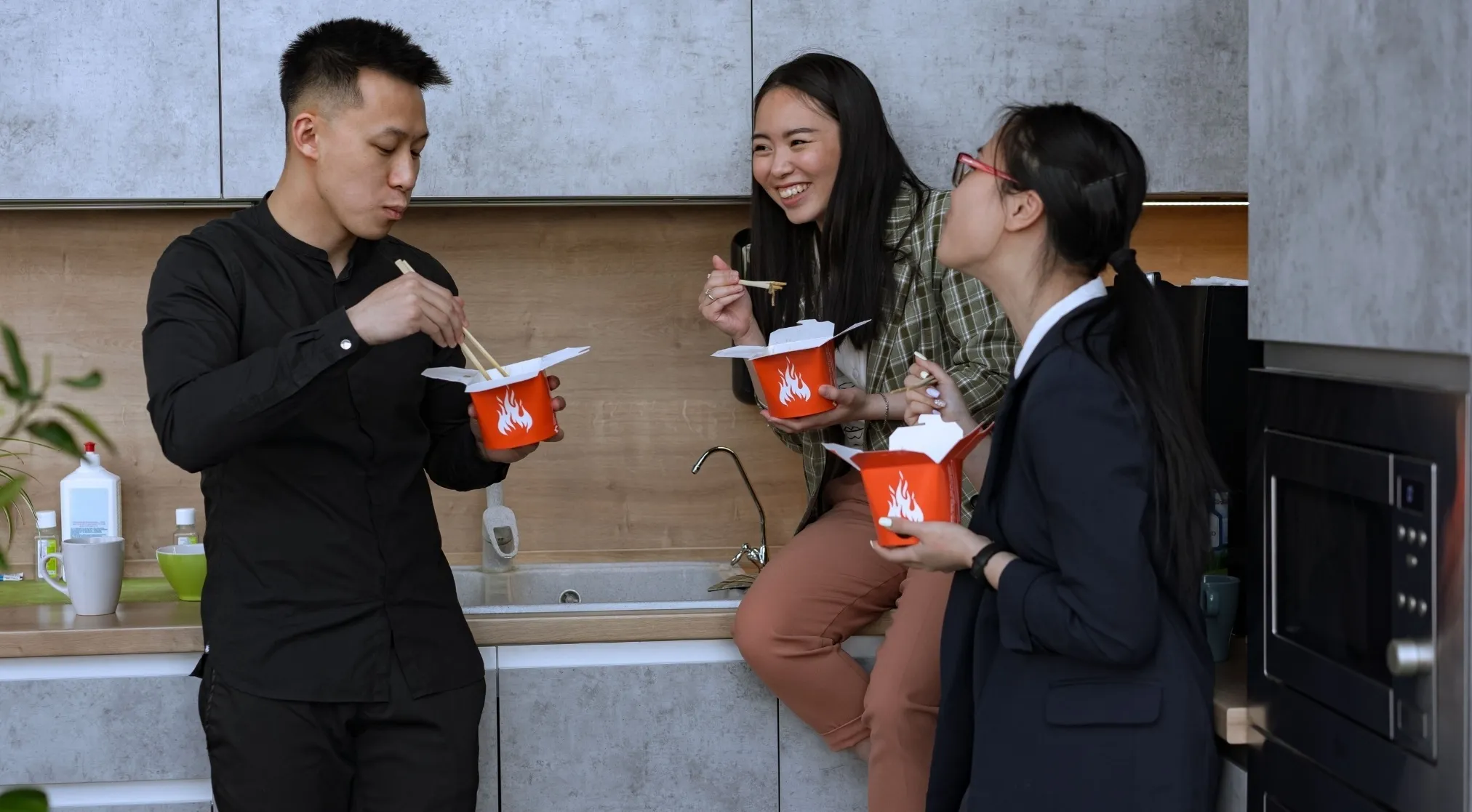 Three colleagues are eating take-out noodles, one man and two women, discussing spelling errors in a casual setting.