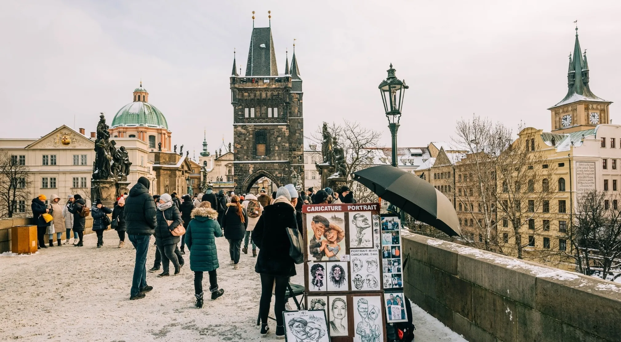 A winter scene on Charles Bridge, Prague, with people walking on snow. A street artist displays caricatures, focusing on spelling error.