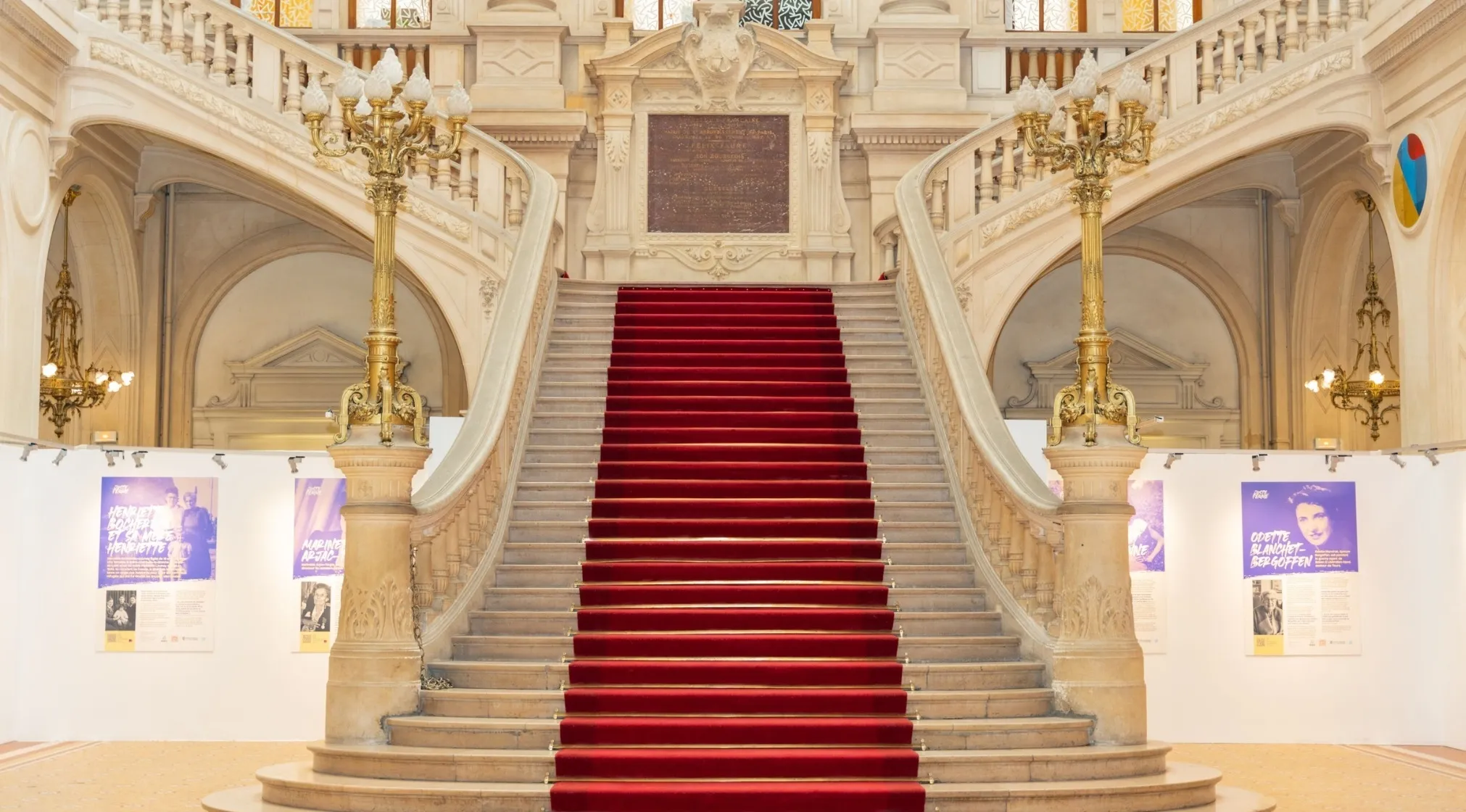 Grand stairway with a red carpet and ornate banisters. Two golden lampposts flank the stairs, adding a touch of glamour.