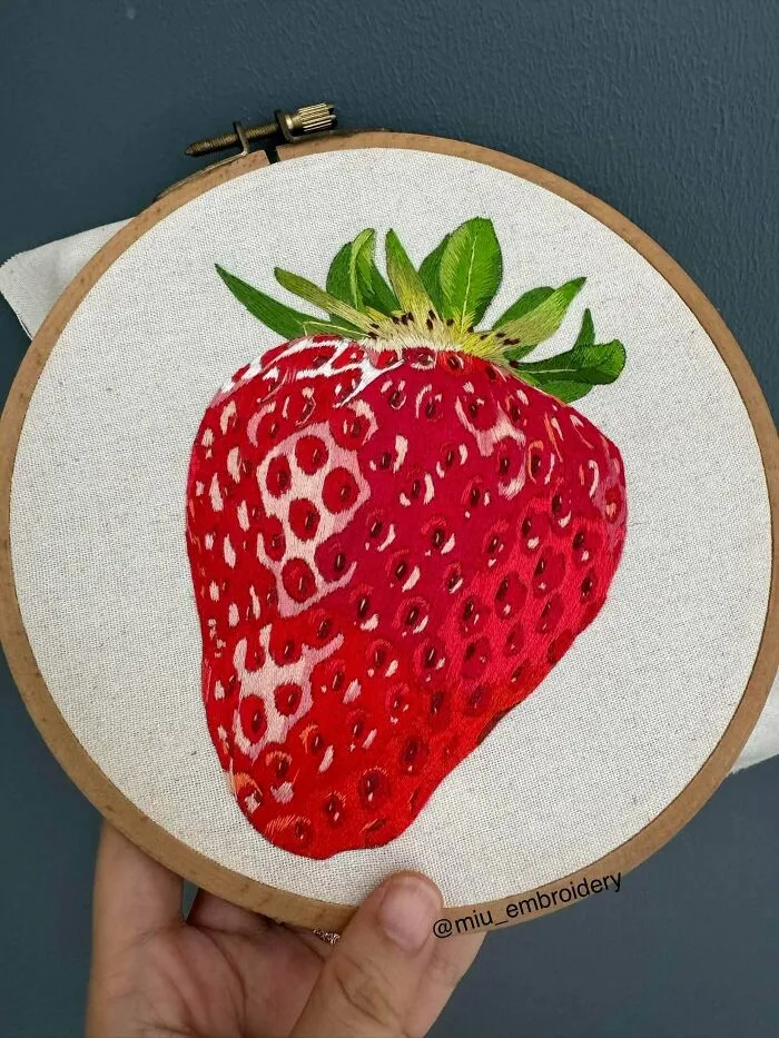 A detailed embroidery design of a large, ripe red strawberry stitched onto fabric inside a wooden hoop