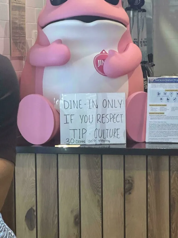A sign on a counter next to a pink frog statue stating Dine-in only if you respect tip culture, emphasizing the need to end tipping.