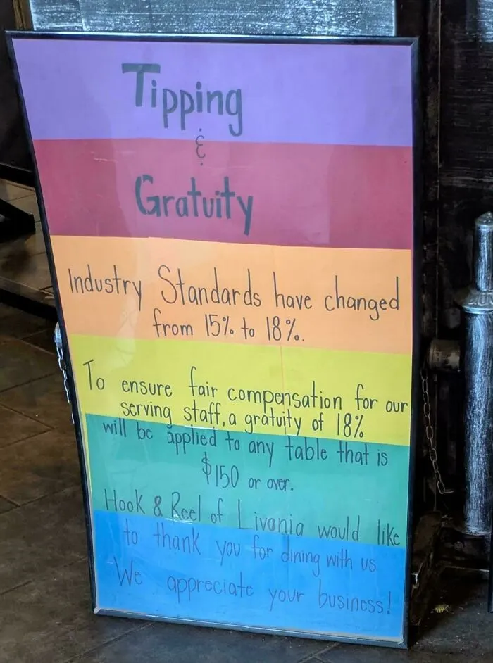 A colorful sign from Hook & Reel of Livonia stating industry standards for tipping and gratuity have changed to 18% for tables over $150, reflecting tipping culture.
