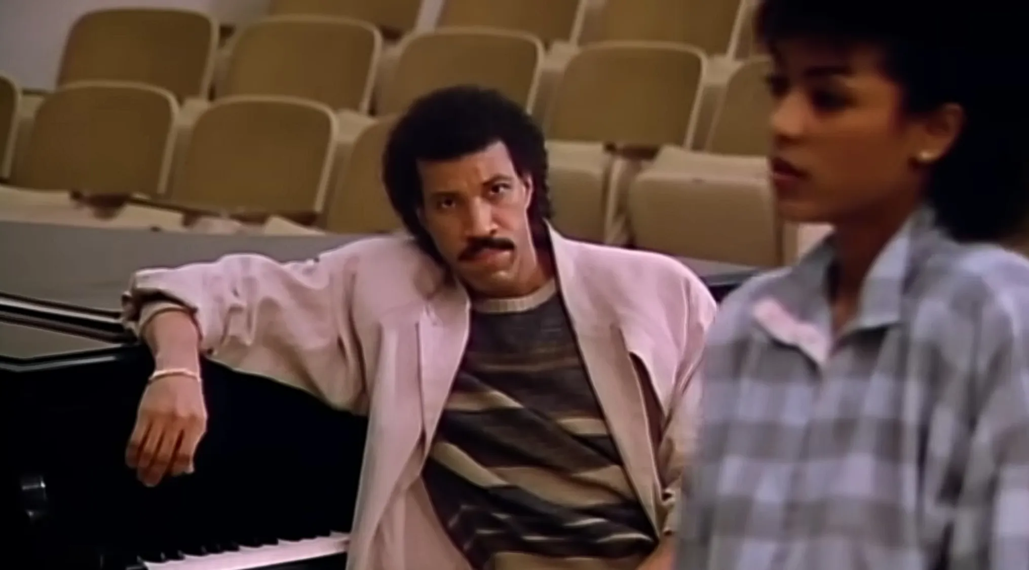 Lionel Richie, from a music video, looking thoughtfully, next to a piano.