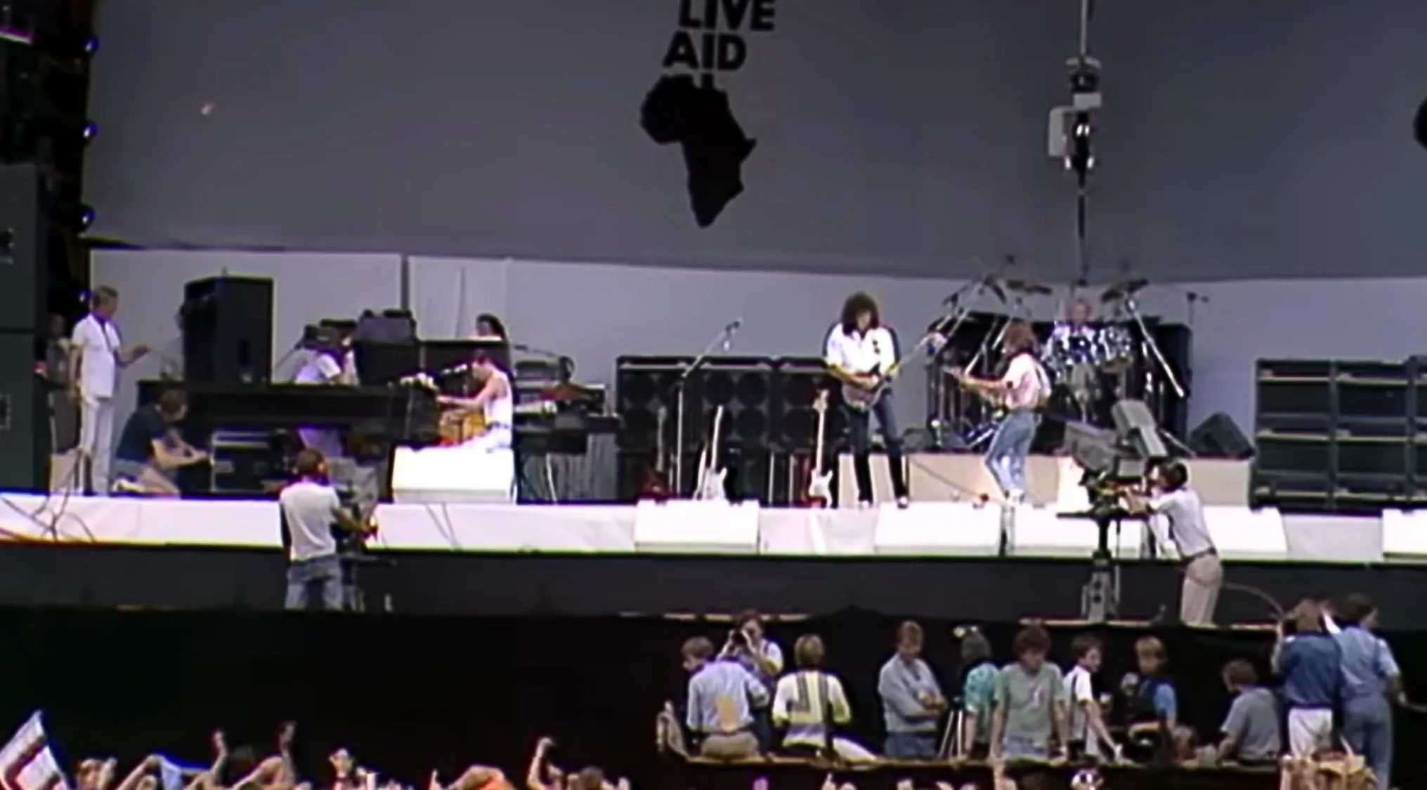 An iconic 80s-90s music band performs live on stage at Live Aid with a large Africa map behind them.