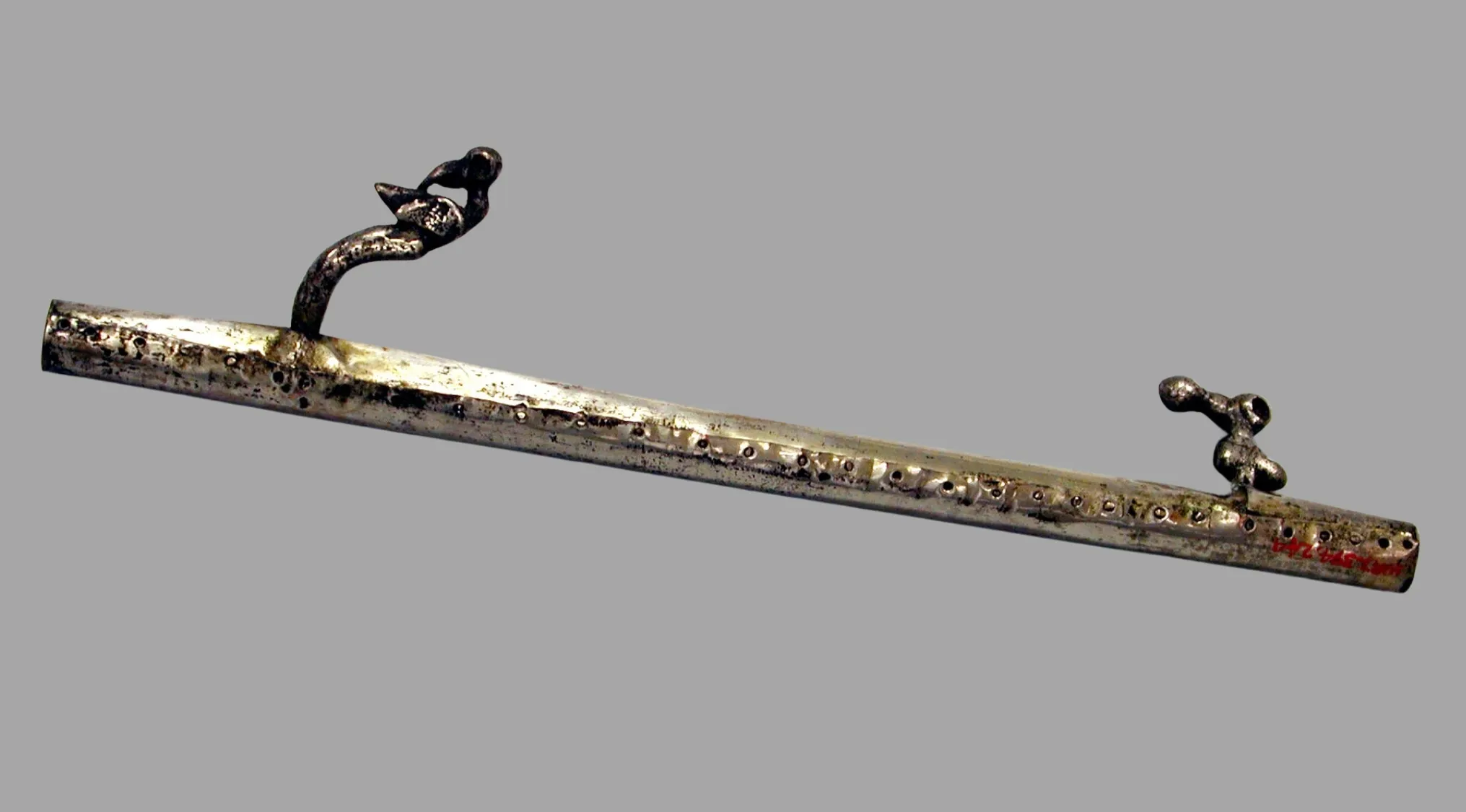 A silver-colored ancient atlatl, likely made of bone or metal, with decorative carvings on a grey background.