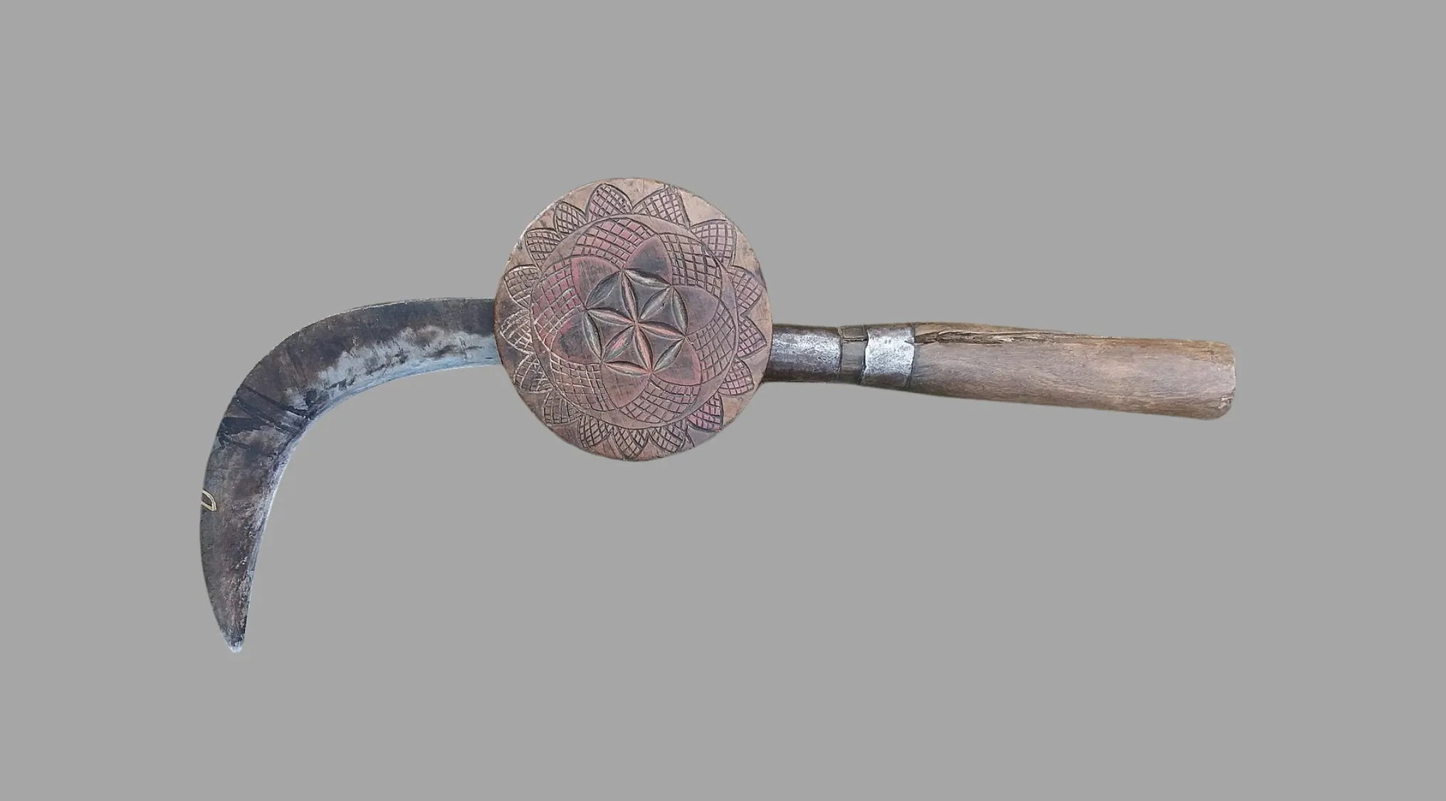 A curved, ancient tool with a metal blade and a wooden handle featuring a circular, carved wooden disc with intricate patterns. What is this artefact?