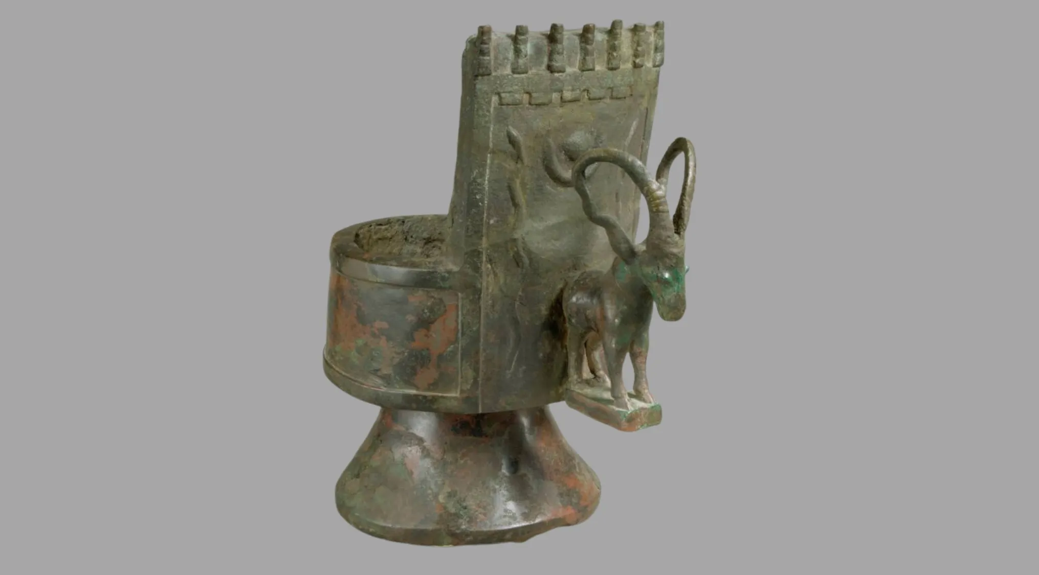 An ancient bronze ceremonial object with a goat figure, showing ornate details and wear. Part of a collection of ancient tools.