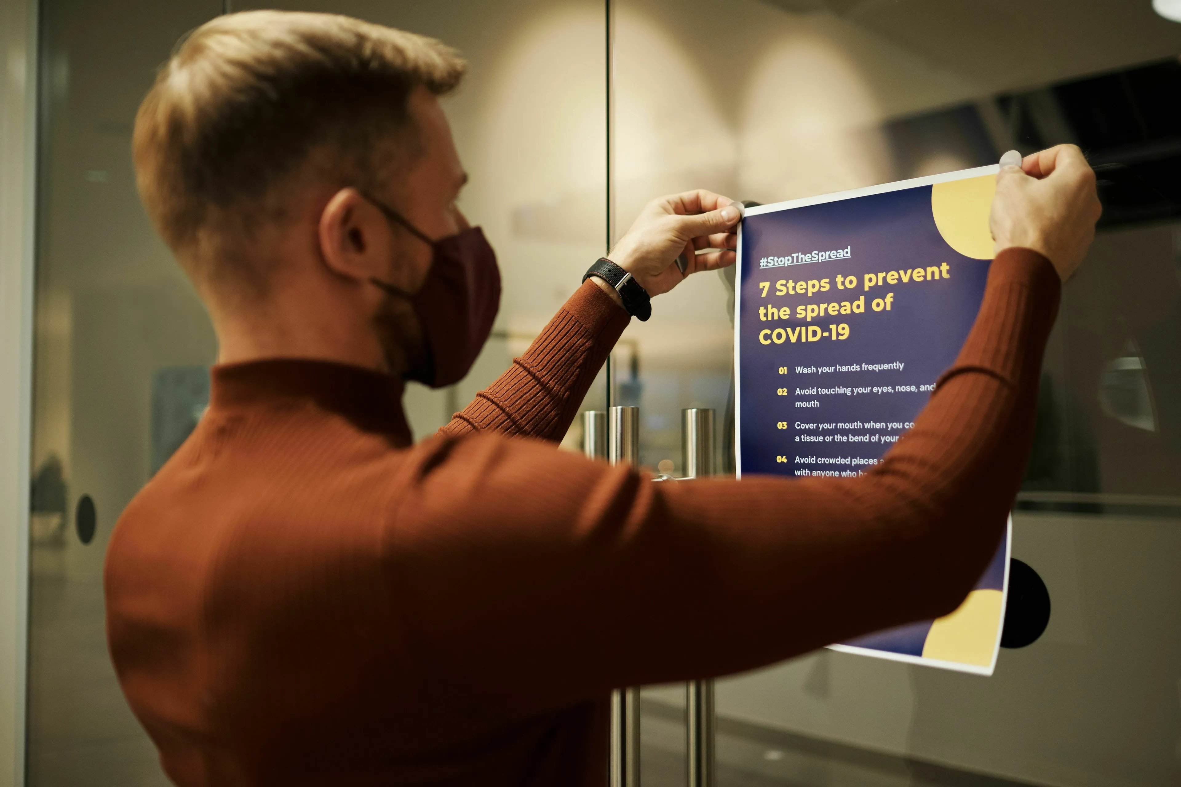 A man in a mask puts up a public health campaign poster with 7 steps to prevent the spread of COVID-19.