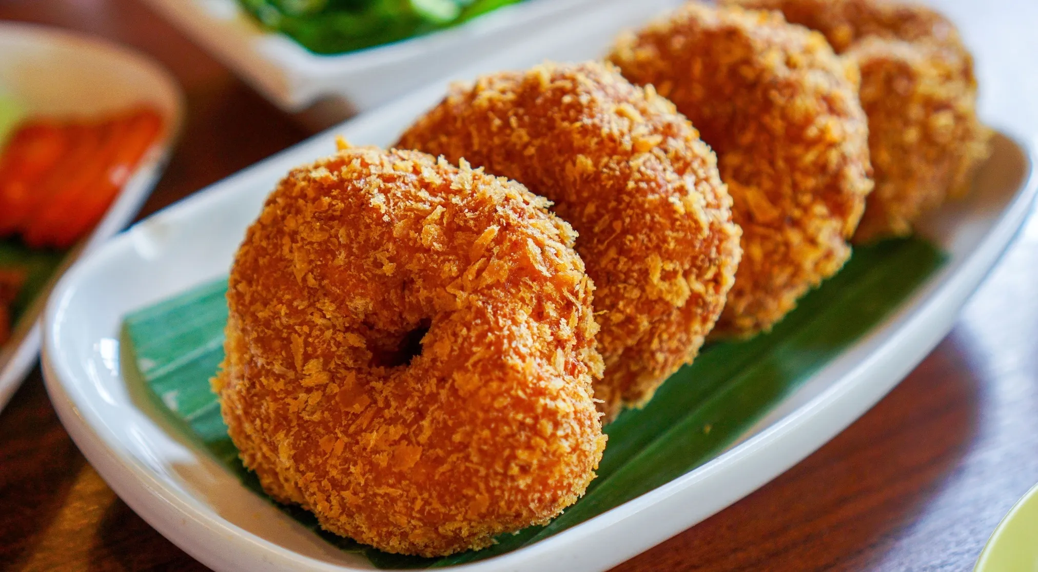 Close-up of three crispy, deep-fried fritters on a white plate with a green banana leaf, perfect example of a small cake.