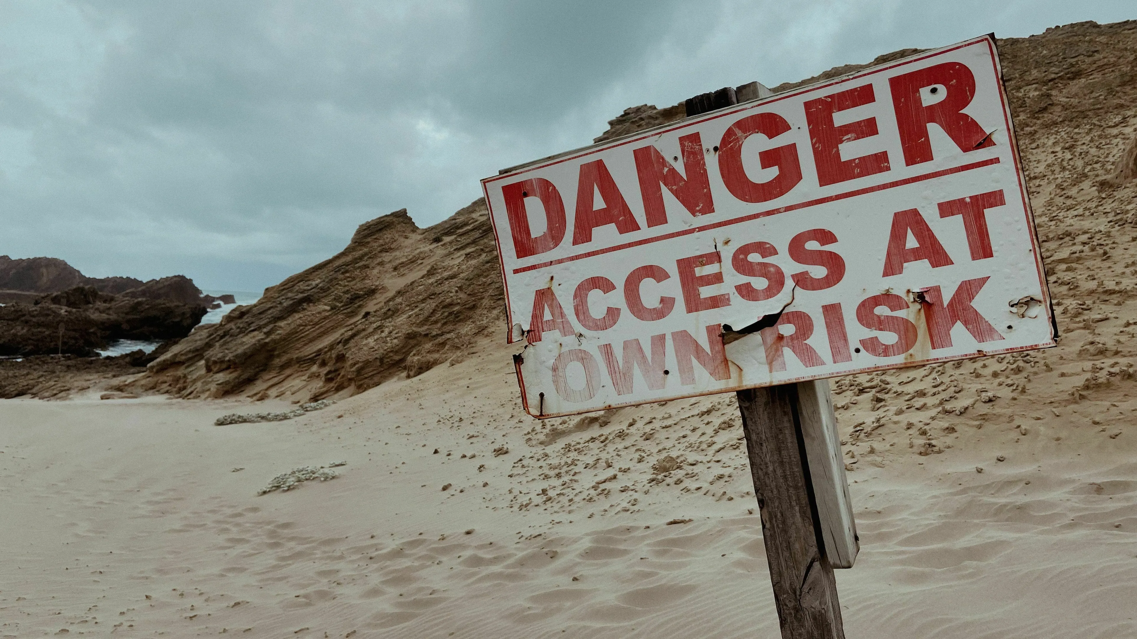 A weathered Danger Access at Own Risk sign on a sandy beach, symbolizing the relationship with risk in Ancient Rome.