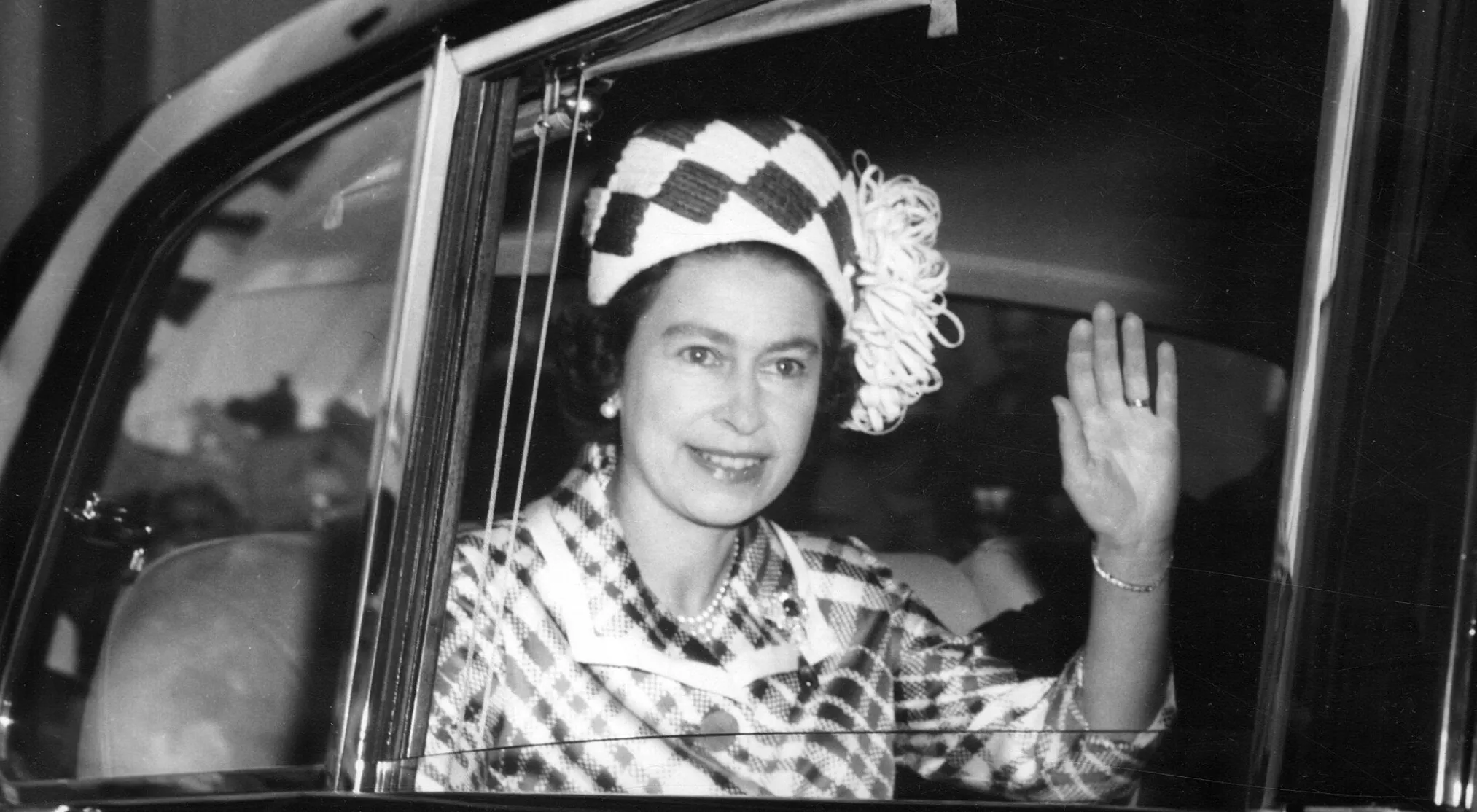 Queen Elizabeth II, smiling and waving from a car window, wearing a patterned hat and pearl necklace. Was she a trained mechanic?