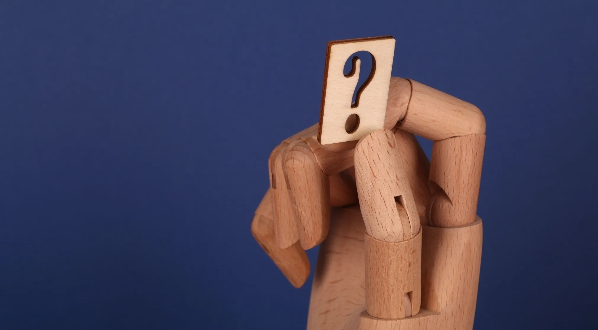 A wooden mannequin hand holds a small wooden cutout of a question mark against a deep blue background. This relates to punctuation.