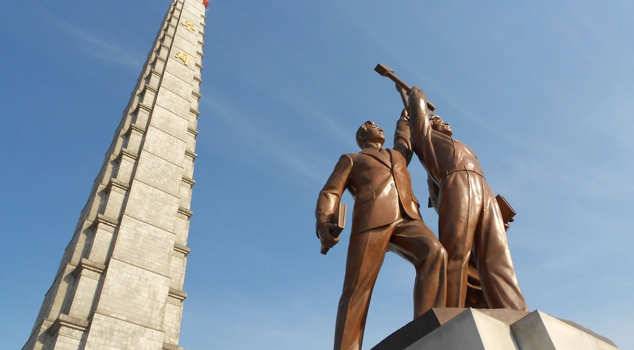 Bronze statues of two men and a tall stone monument against a blue sky, representing China's socialist system.