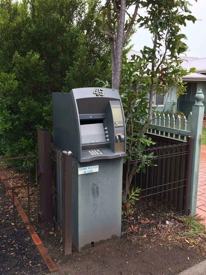 An outdoor ATM next to a gate and trees, representing old junk possibly repurposed for a new use.